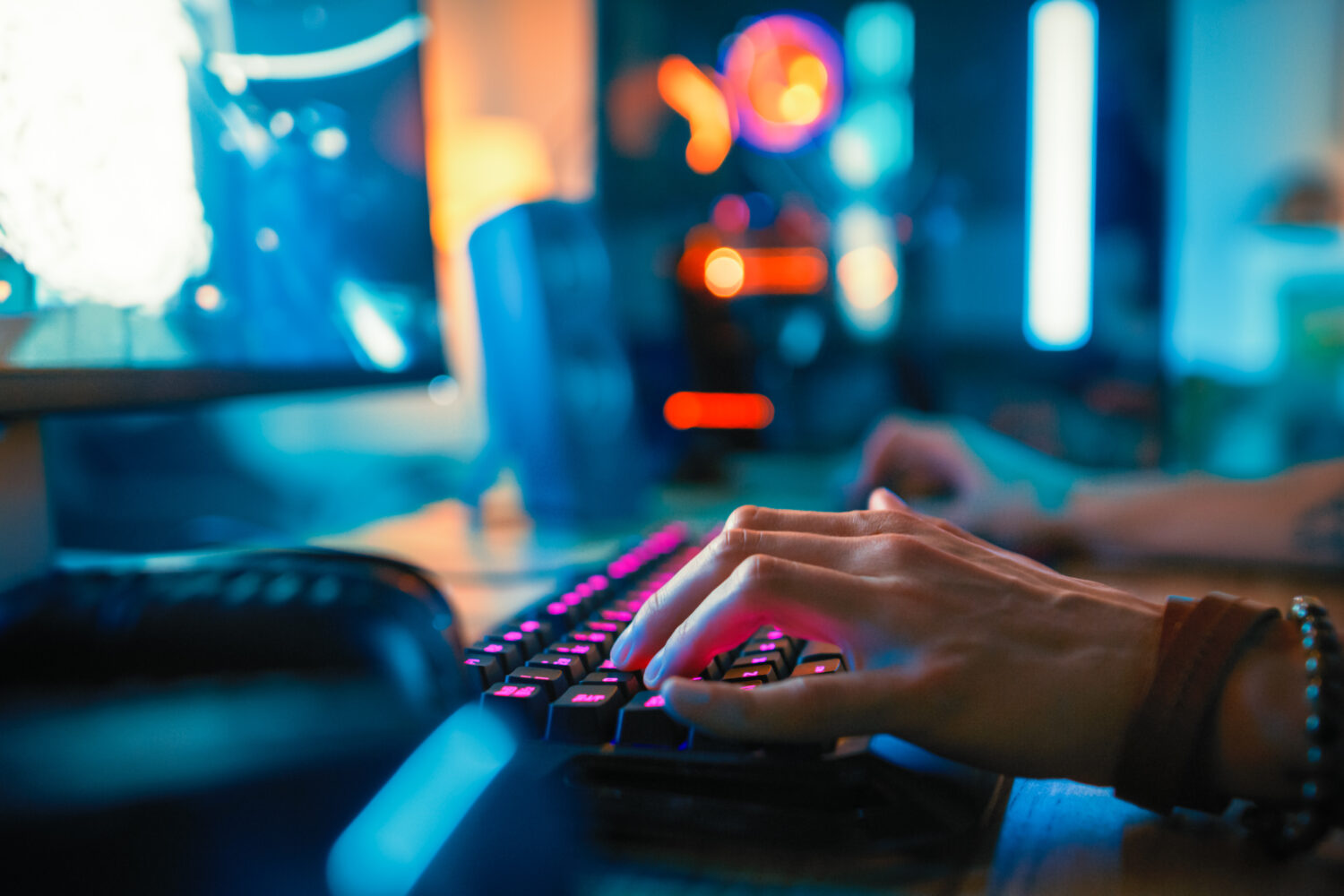 Close-Up Hands Shot Showing a Gamer Using the Keyboard while Playing an Online Shooter Video Game. Keyboard has Pink Neon Lights in Buttons. Gamer is Wearing a Bracelet. Room is Dark.