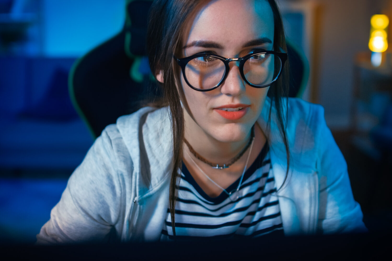Excited and Pretty Gamer Girl in Glasses is Playing Online Video Game on Her Personal Computer. Room and PC have Colorful Warm Neon Led Lights. Cozy Evening at Home.