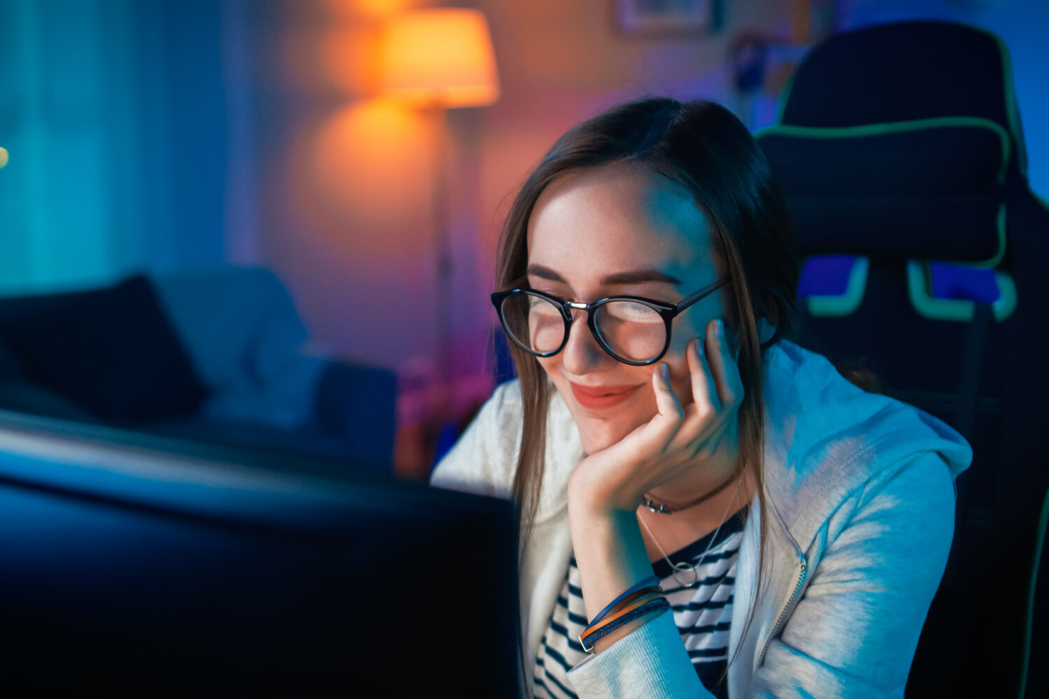 Beautiful Happy and Sentimental Young Girl Blogger Watching Videos on a Computer. She Has Dark Hair and Wears Glasses. Screen Adds Reflections to Her Face. Cozy Room is Lit with Warm Light.