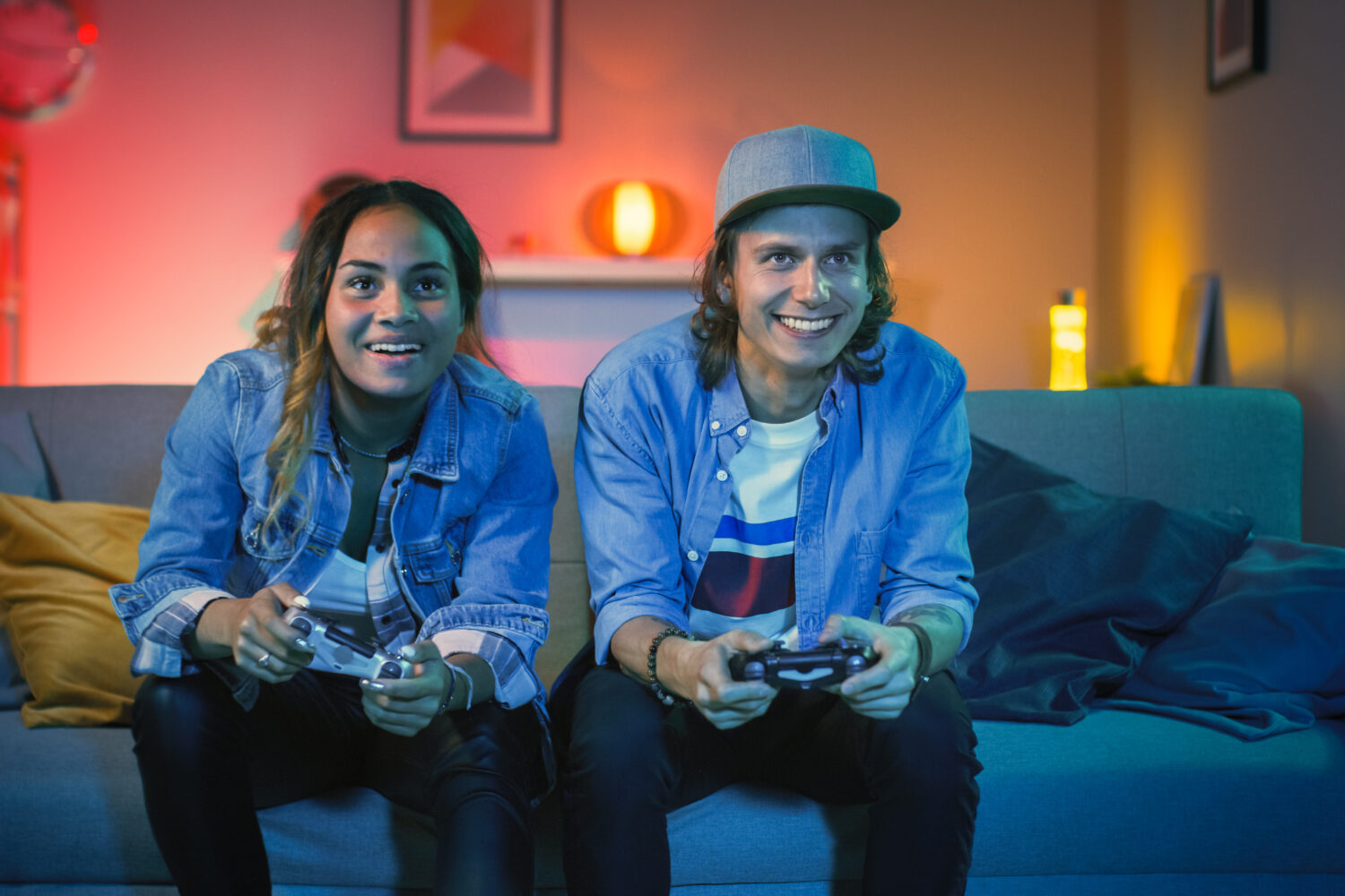 Excited Black Gamer Girl and Young Man Sitting on a Couch and Playing Video Games on Console. They Plays with Wireless Controllers. Cozy Room is Lit with Warm and Neon Light.