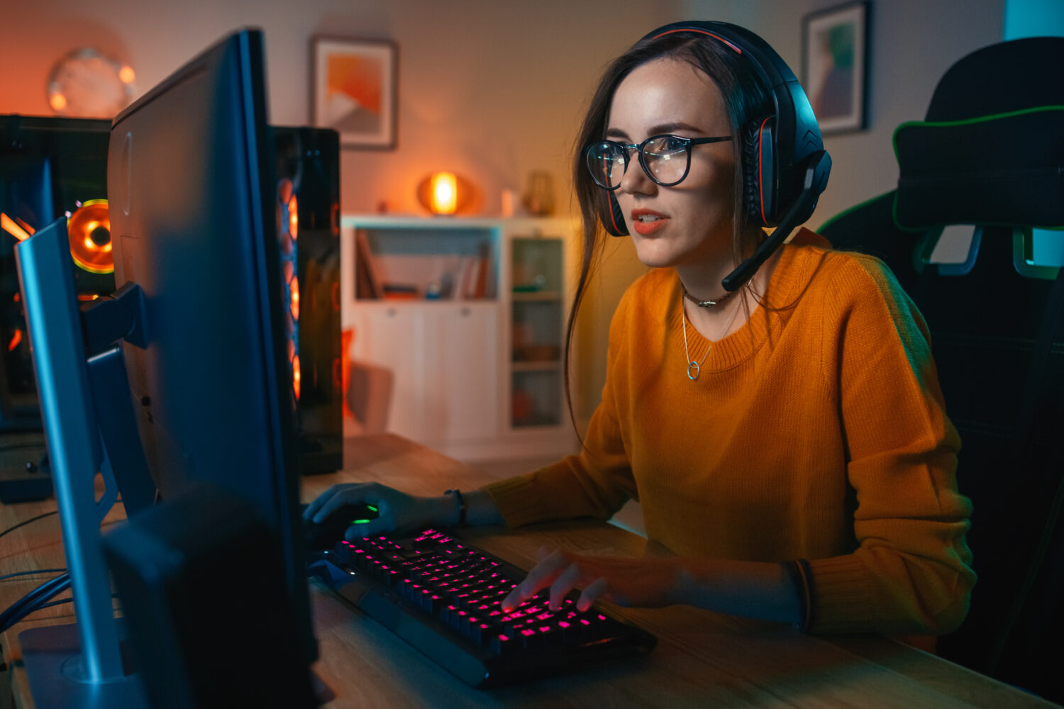 Excited Gamer Girl in Headset with a Mic Playing Online Video Game on Her Personal Computer. She Talks to Other Players. Room and PC have Colorful Warm Neon Led Lights. Cozy Evening at Home.